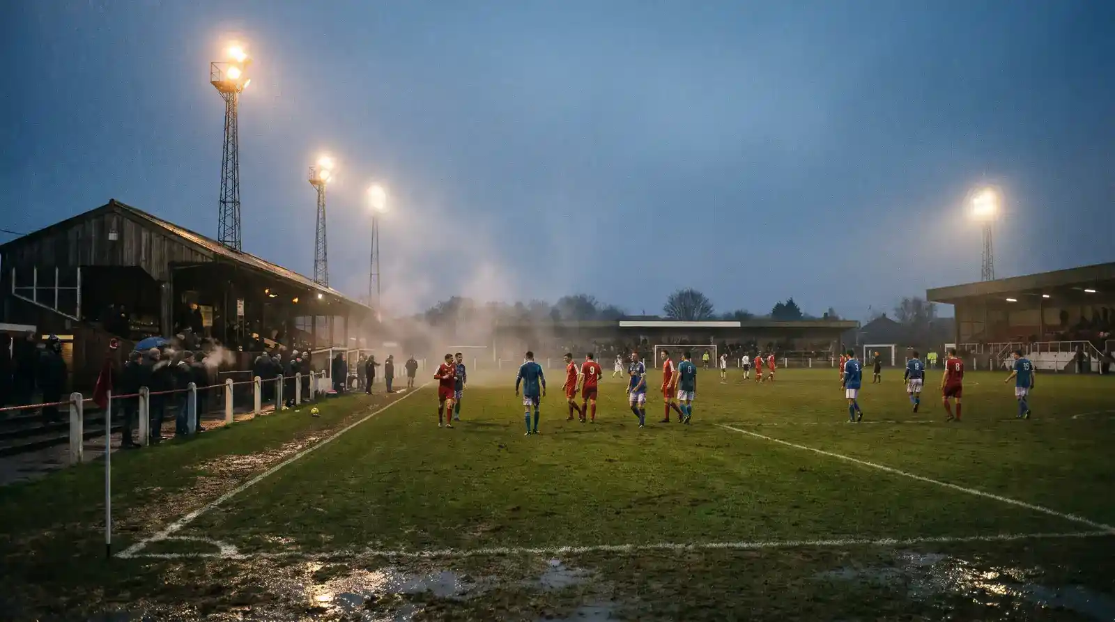 Floodlit non-league English football stadium at dusk with a sparse crowd and players warming up on a grass pitch